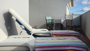 Un balcón con dos camas a rayas, una mesa con una botella y un vaso, y una silla.