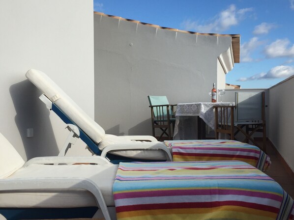 Un balcón con dos camas a rayas, una mesa con una botella y un vaso, y una silla.