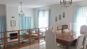 Un comedor con una mesa de madera y sillas blancas, una araña y vistas a un balcón con cortinas azules.