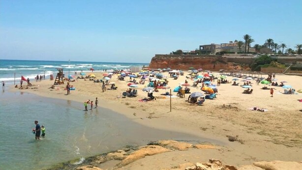 Une plage bondée où les gens se prélassent au soleil et se baignent, une tour de surveillance des sauveteurs et des bâtiments en arrière-plan.
