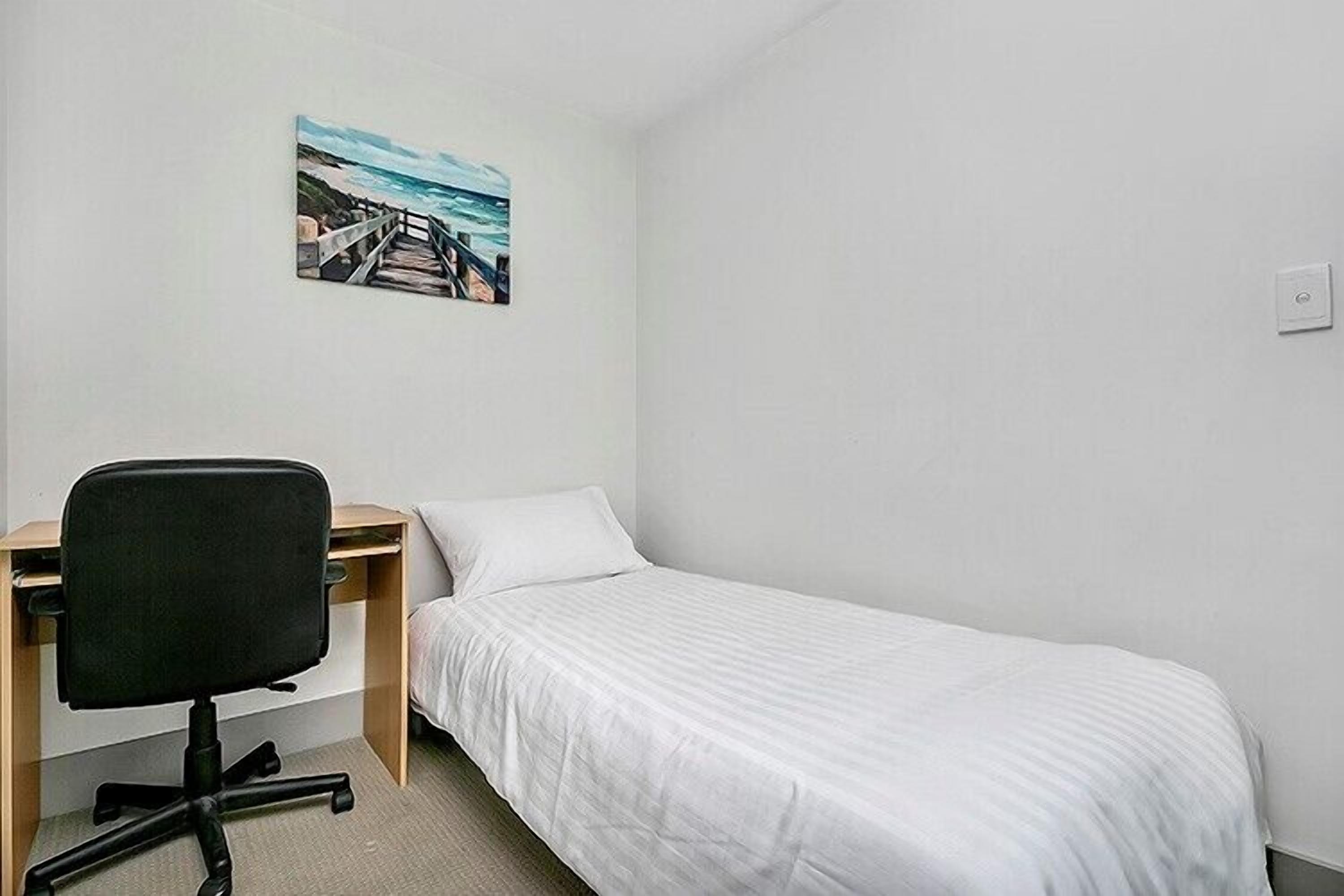 Una habitación pequeña y limpia con una cama individual, un escritorio y una silla. Hay un cuadro en la pared que representa una escena costera.