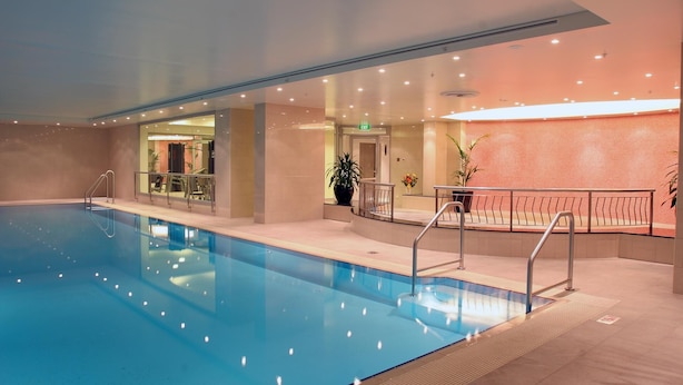 Ein Hallenschwimmbad mit klarem Wasser, umgeben von Fliesenboden und -wänden. Glastüren führen zu einem Raum mit Trainingsgeräten.