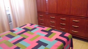 Quarto com uma cama coberta por um edredom colorido com padrão geométrico, cômoda de madeira com várias gavetas e um armário com porta visível.