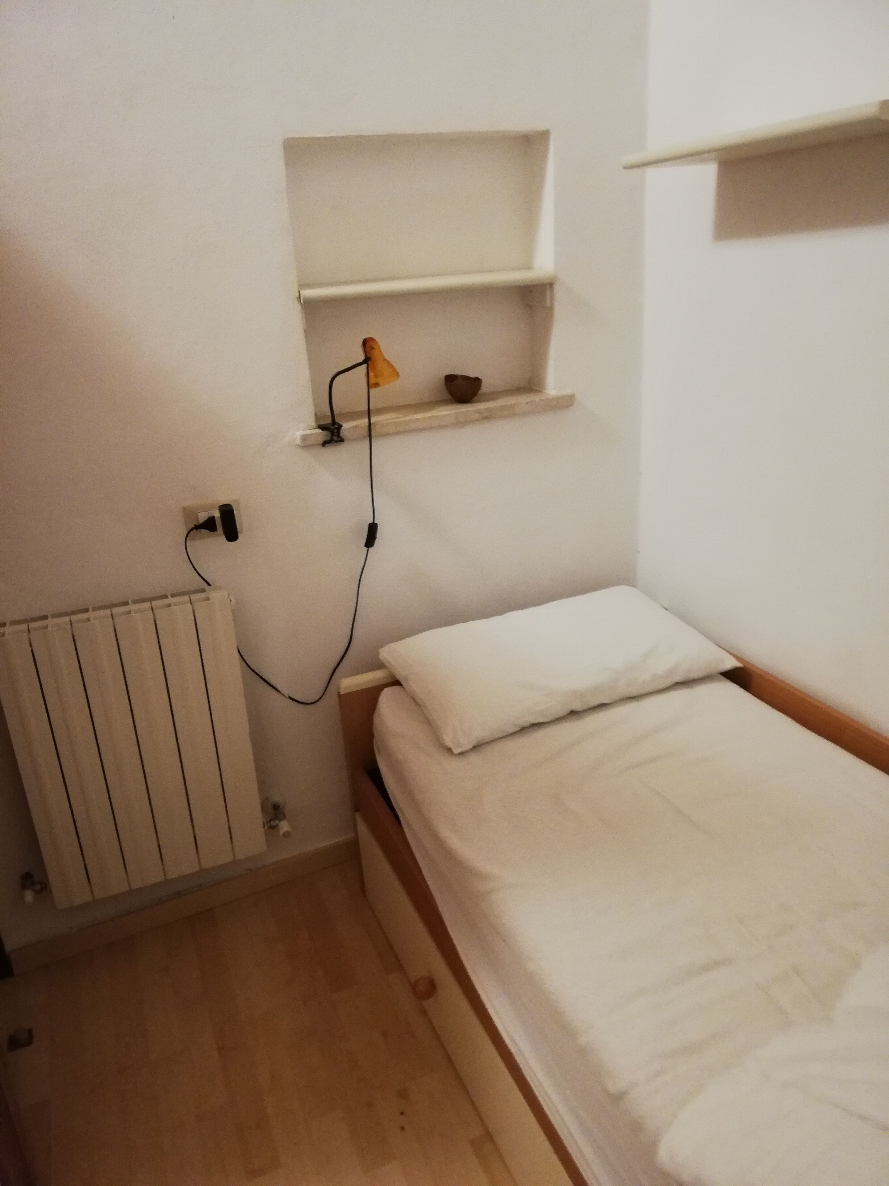 Une petite pièce simple, avec un lit, une étagère sur laquelle se trouve une lampe et un radiateur.