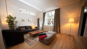 Un salon moderne comprenant un canapé, un pouf et une table basse, une grande fenêtre avec des rideaux et un lampadaire.
