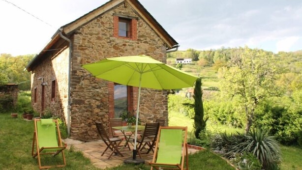 A stone house with a patio, two green chairs, and a large umbrella. - Une Sorcière sur le toit - Gîte** en Aveyron: Countryside and absolute tranquillity! (Firmi)