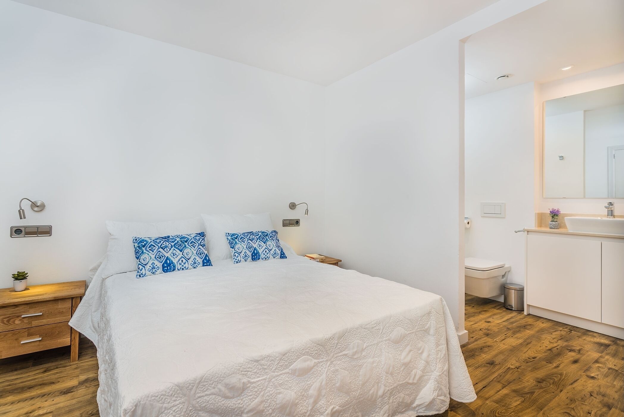 A bedroom with a bed, bedside tables, a wooden floor, a white wall, and a bathroom with a sink and toilet.