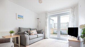 A modern living room with a grey sofa, a small table, a TV, and a view of the outside. - Lavender - Two Bedroom Cottage, Sleeps 4 (Wadebridge)