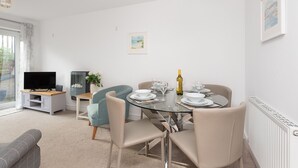A modern living room with a dining area, featuring a glass table, beige chairs, a flat-screen TV, and a small side table with a plant. - Lavender - Two Bedroom Cottage, Sleeps 4 (Wadebridge)