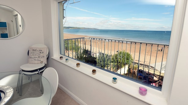 A room with a large window overlooking a beach, a round mirror, a white chair, and a small table.