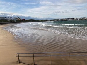 Une plage avec une rambarde métallique au premier plan, des vagues qui viennent doucement lécher le rivage, et une vue lointaine des bâtiments qui bordent la côte.
