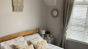Een net opgemaakt bed met een houten hoofdbord, een nachtkastje met een lamp, een wanddecoratie en een raam met gordijnen.