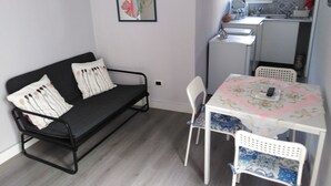 Et lille, moderne rum med en sort sofa, et hvidt bord med stole, et tekøkken med vask og komfur, samt et billede i ramme på væggen.