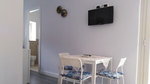 Et lille spiseområde med et hvidt bord og stole, et fladskærms-tv ophængt på væggen og en dør til et badeværelse.