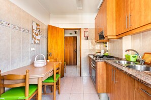 Uma cozinha compacta com armários de madeira, fogão e pia. Há uma mesa de jantar com cadeiras e um quadro de avisos na parede.