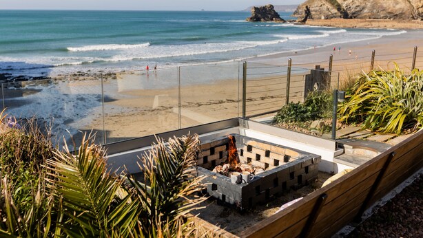 A beach view with a fire pit, wooden deck, and a cliff in the background.