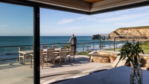 A patio with outdoor furniture overlooking the ocean, a person walking, and a cliff in the background.