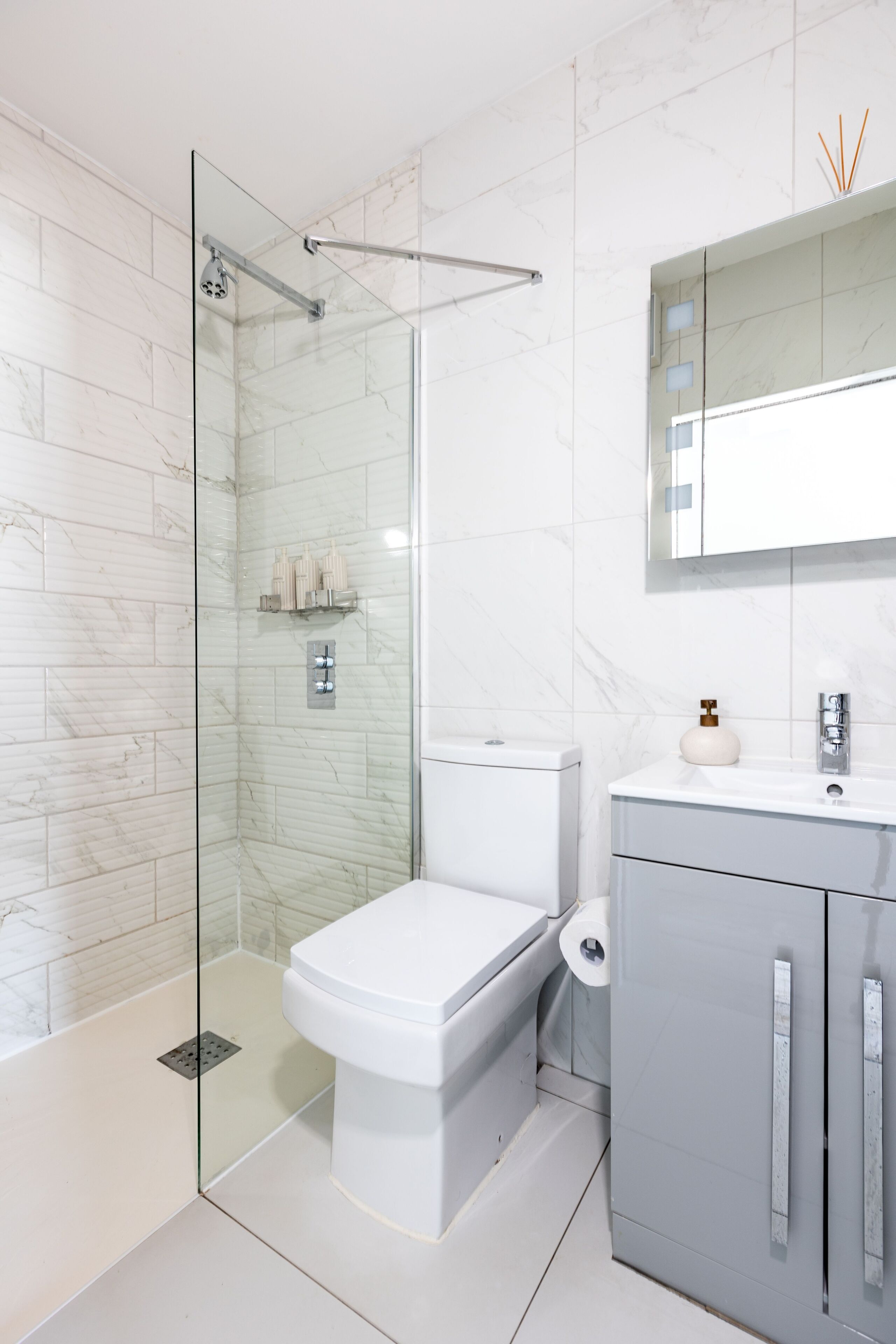 Un baño moderno con ducha envuelta en cristal, inodoro blanco, mueble gris y un espejo sobre el lavabo.