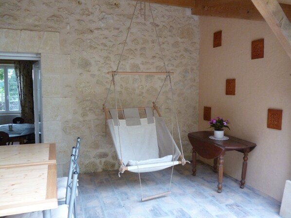 Una habitación rústica con pared de piedra, techo de madera, un sillón colgante, una pequeña mesa de madera con una planta en maceta y una ventana con cortinas.