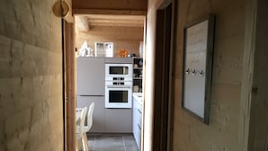 Een smalle houten gang die leidt naar een keuken met een witte koelkast, oven en een wandlamp.