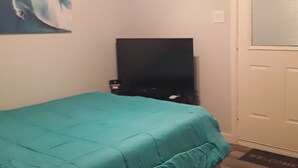 Un dormitorio con colcha color turquesa, ventilador de techo, televisor sobre un soporte y puerta con ventanal.