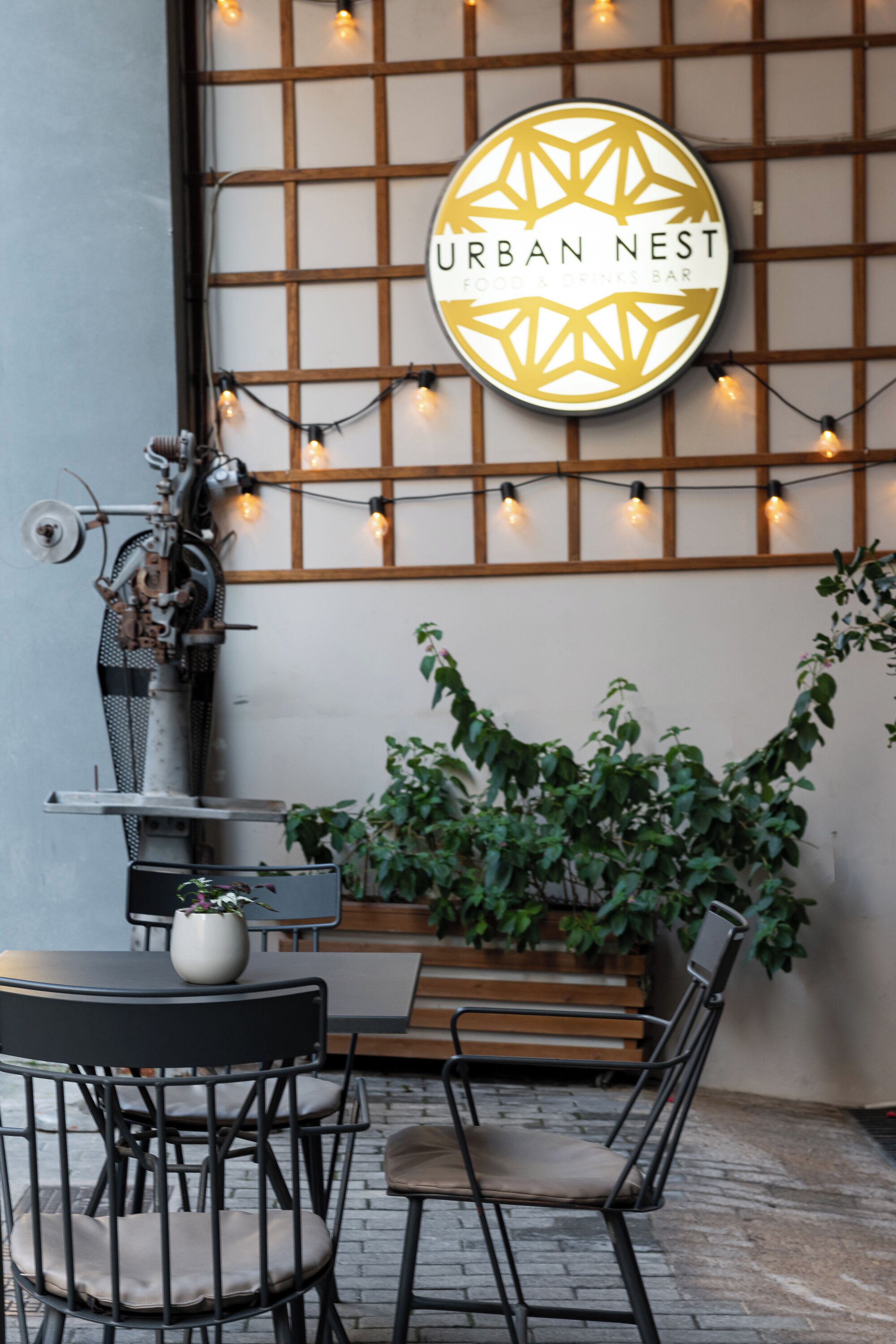 An outdoor seating area with a table and chairs, a hanging sign for 'URBAN NEST Food & Drinks Bar', and a potted plant.
