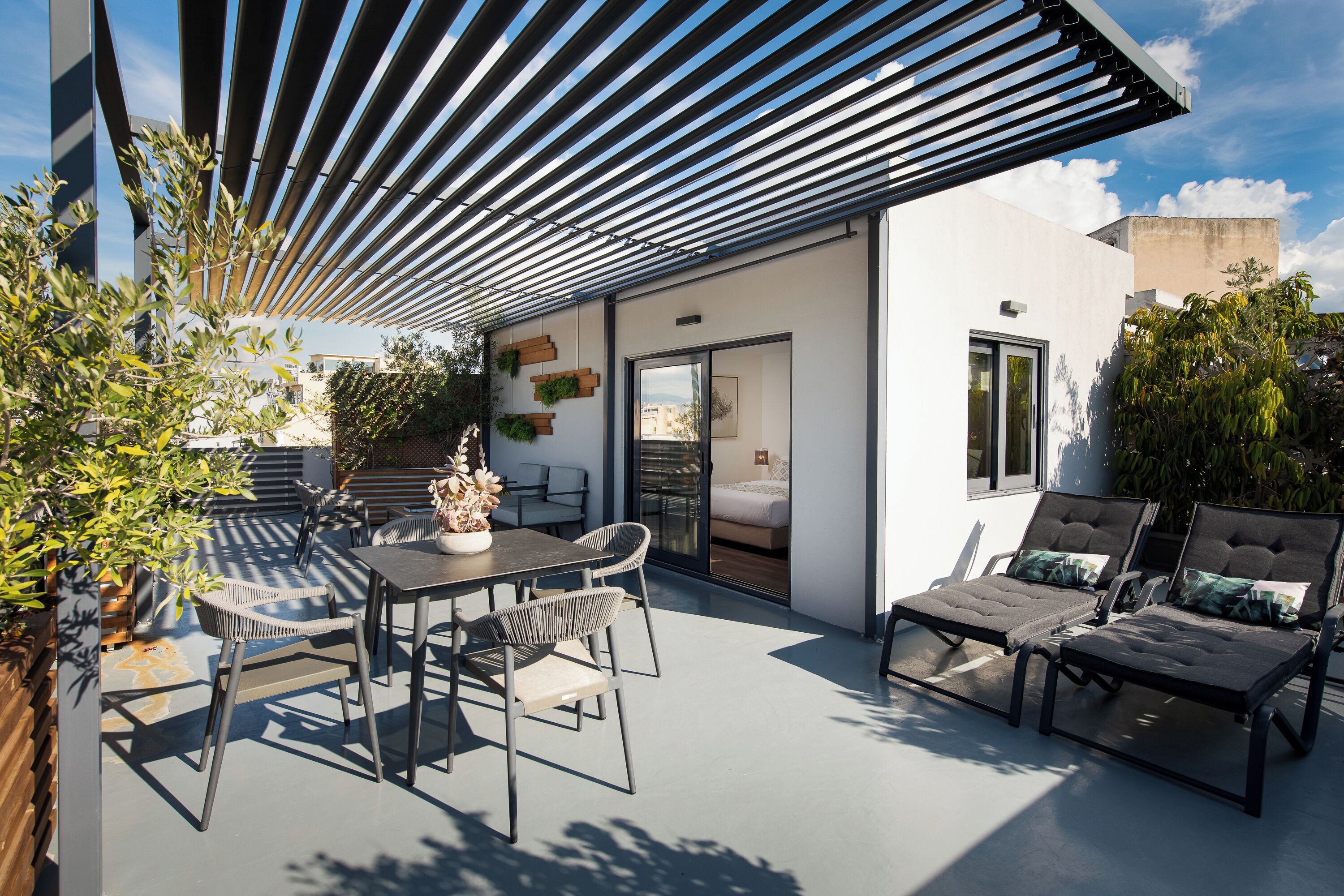 Une terrasse extérieure moderne, dotée d’une table et de chaises, d’un espace salon abrité et offrant une vue sur l’intérieur grâce à une porte vitrée.