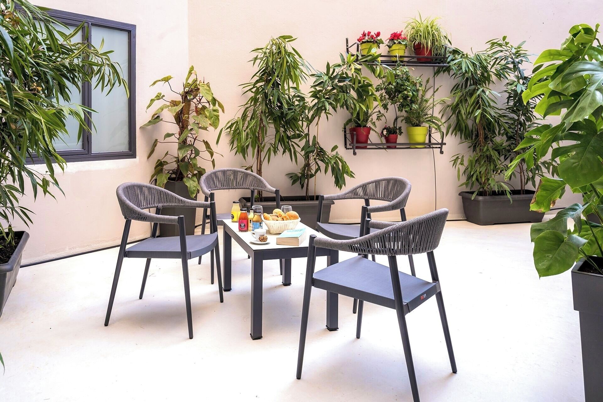 A patio with four grey wicker chairs and a table set for breakfast. There are various potted plants on shelves and hanging pots around the area.