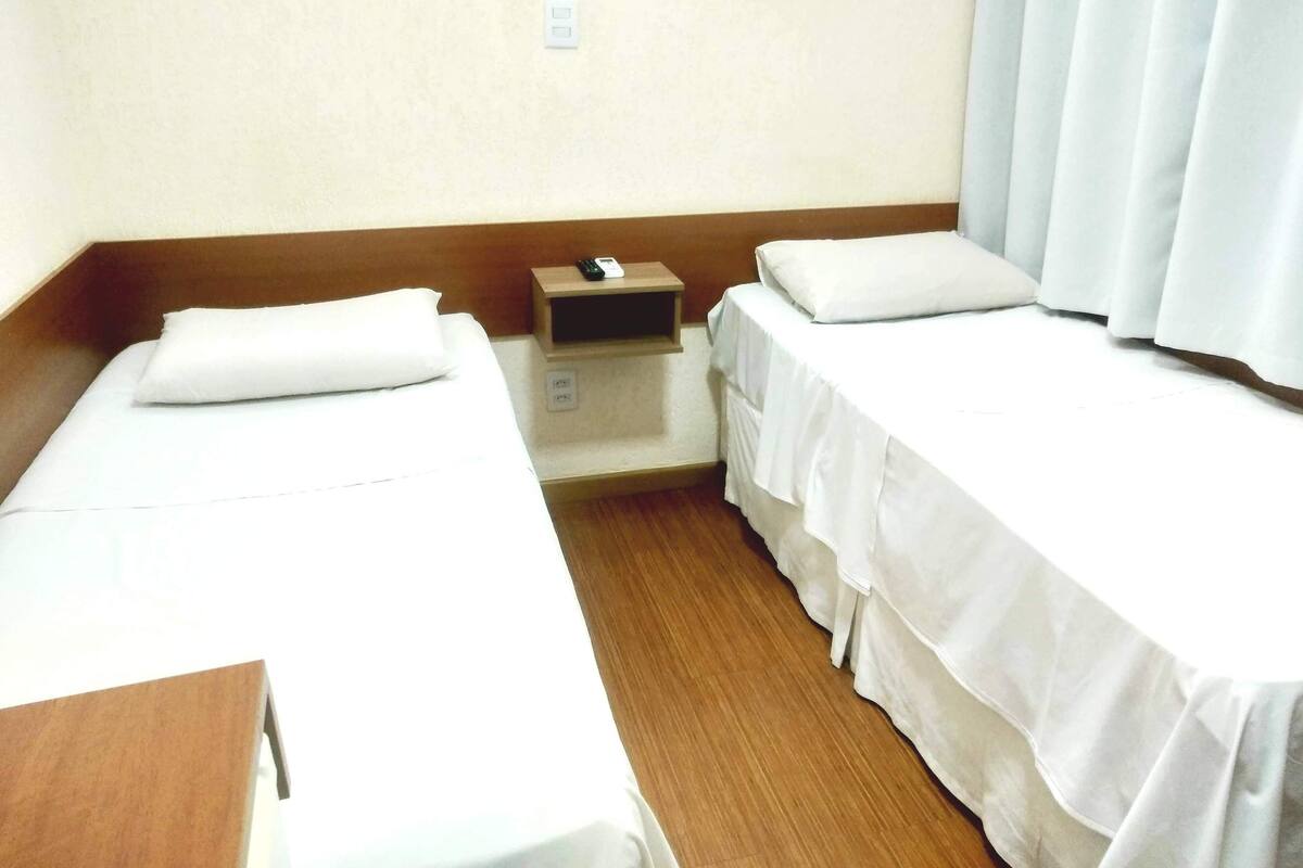 Una habitación de hotel con dos camas individuales, una mesita de noche de madera y una cortina blanca.