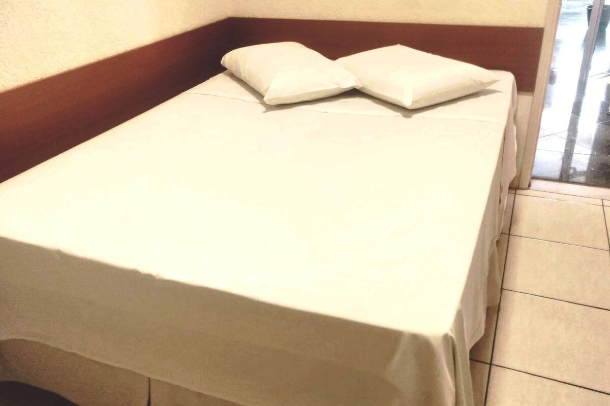 Una cama pequeña y bien hecha con sábana blanca y dos almohadas en una habitación lateral con cabecera de madera y ventana.
