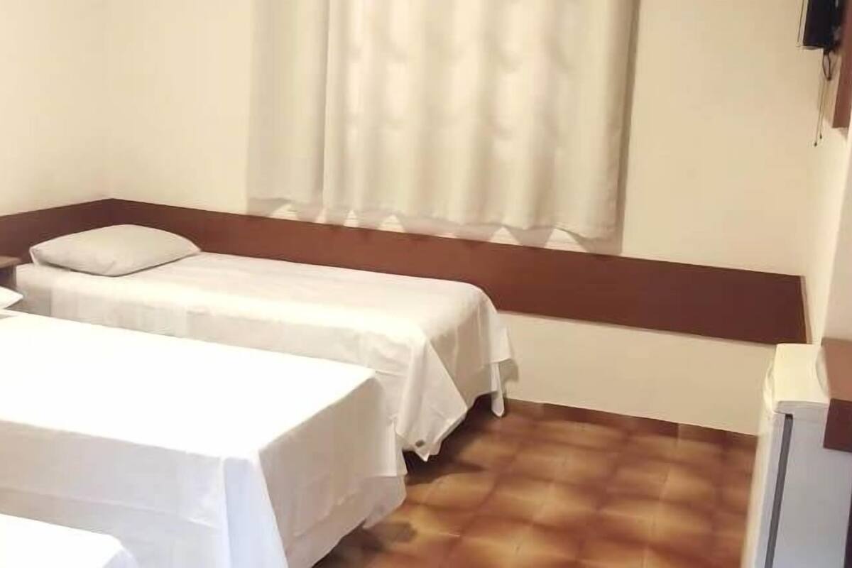Una habitación pequeña y sencilla con dos camas, una mesita de noche, una ventana con cortinas y un banco de madera.