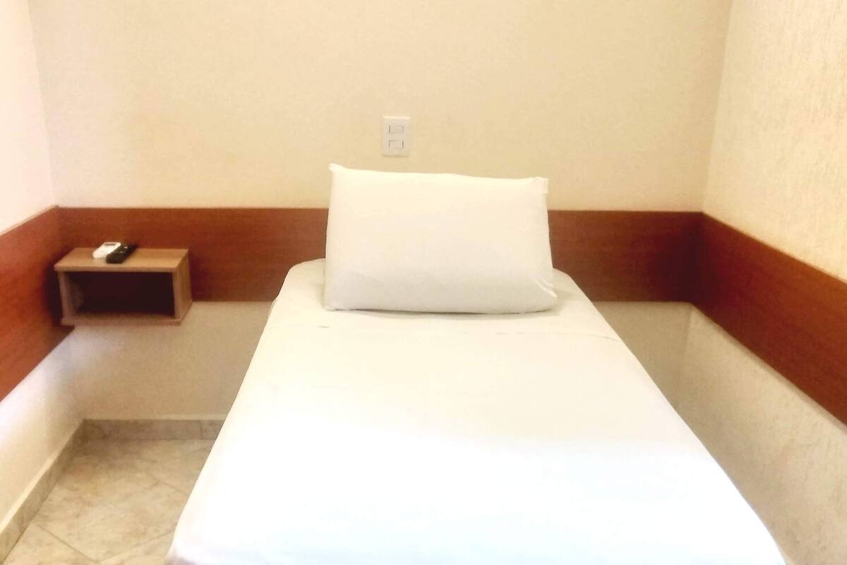 Una habitación pequeña con cama individual, una colcha blanca, una almohada y una mesita de noche con un control remoto.