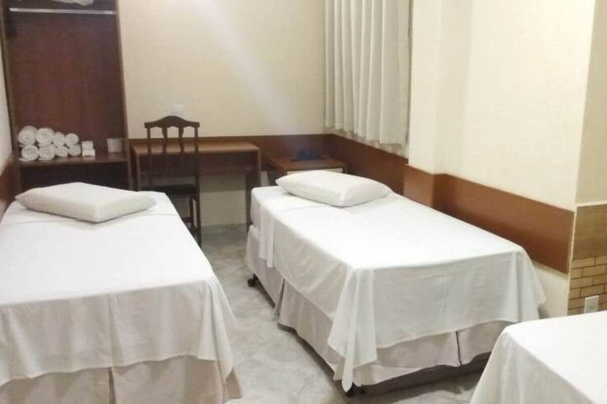 Habitación de hotel con dos camas individuales, un armario de madera, una mesita pequeña y una ventana con cortinas.