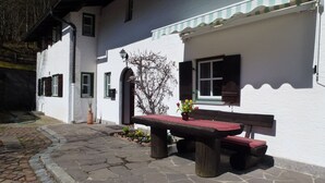 A white building with a stone-paved courtyard, a wooden bench, and a small table with a vase of flowers. - Haus Waldklause (Schoenau am Koenigssee)