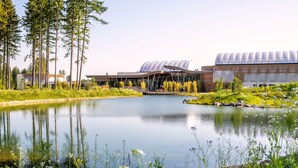 A modern building with a unique curved roof, a small bridge, and a pond in the foreground. - Center Parcs Bungalowpark Allgäu (Leutkirch im Allgau)