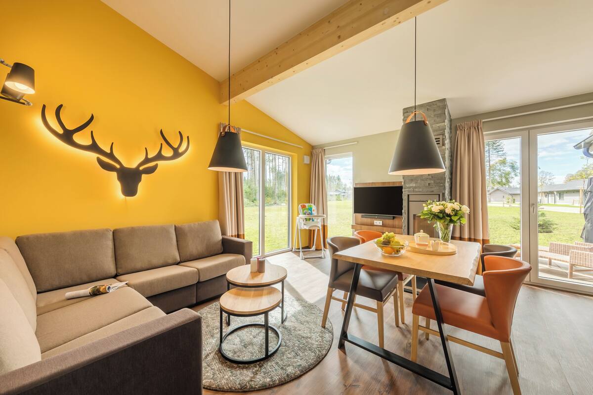 A modern living room with a yellow accent wall, a mounted deer head, a sofa, a dining table with chairs, and a TV.