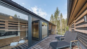 A modern wooden deck with two lounge chairs, a glass door leading to a room, and a view of trees. - Center Parcs Bungalowpark Allgäu (Leutkirch im Allgau)