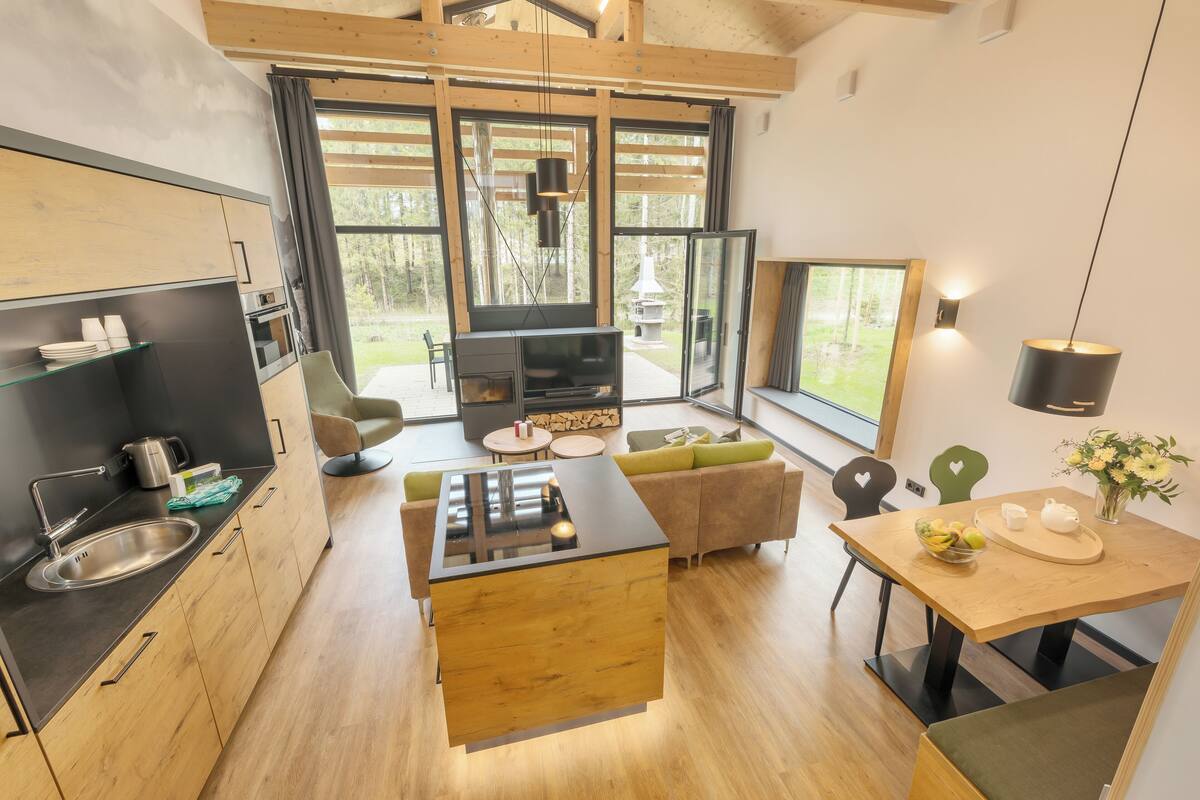 A modern kitchen with a central island, a dining area with a wooden table, and large windows offering a view of the outdoors.