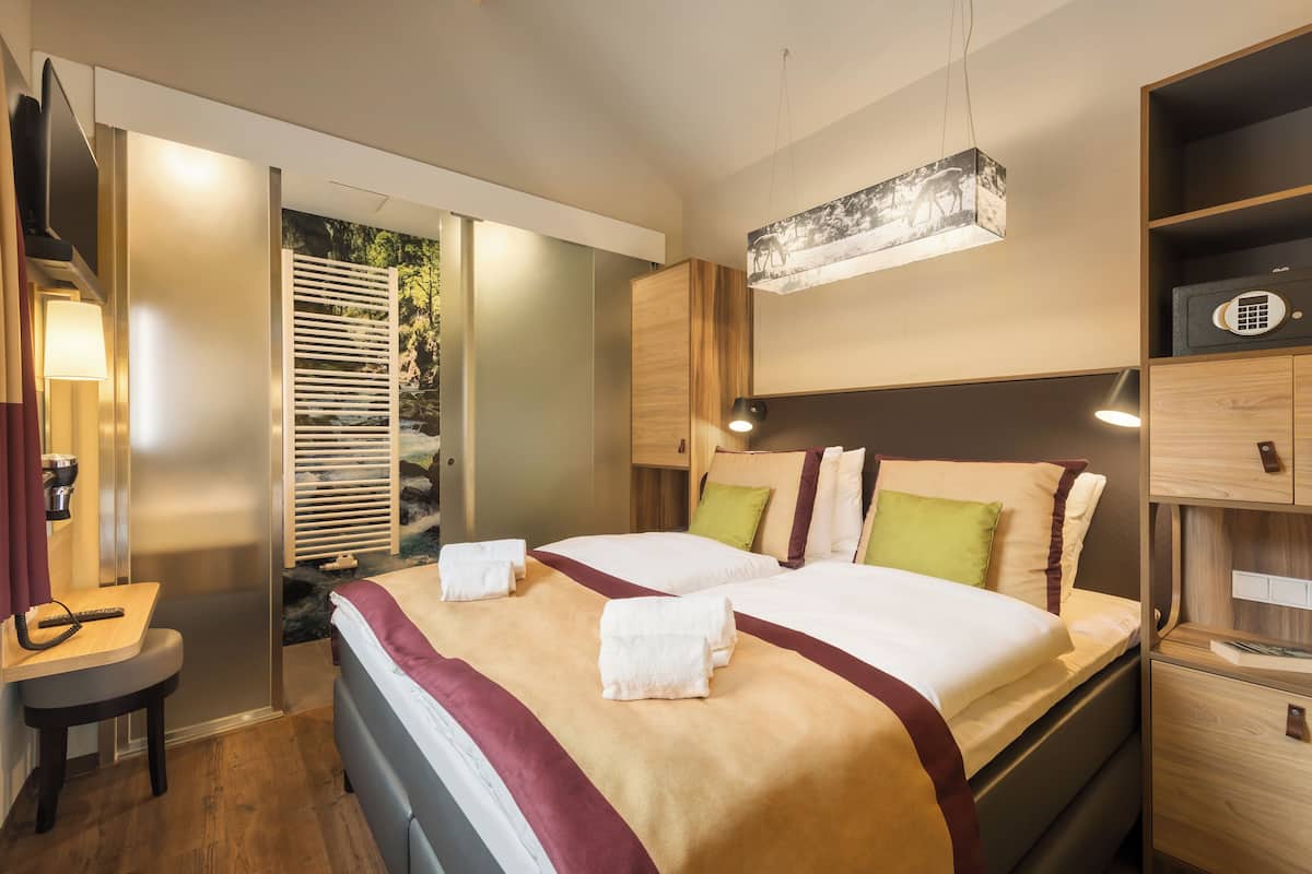 A modern hotel room with a large bed, wooden headboard, and a built-in TV. There is a desk with a lamp, a wardrobe, and a safe.