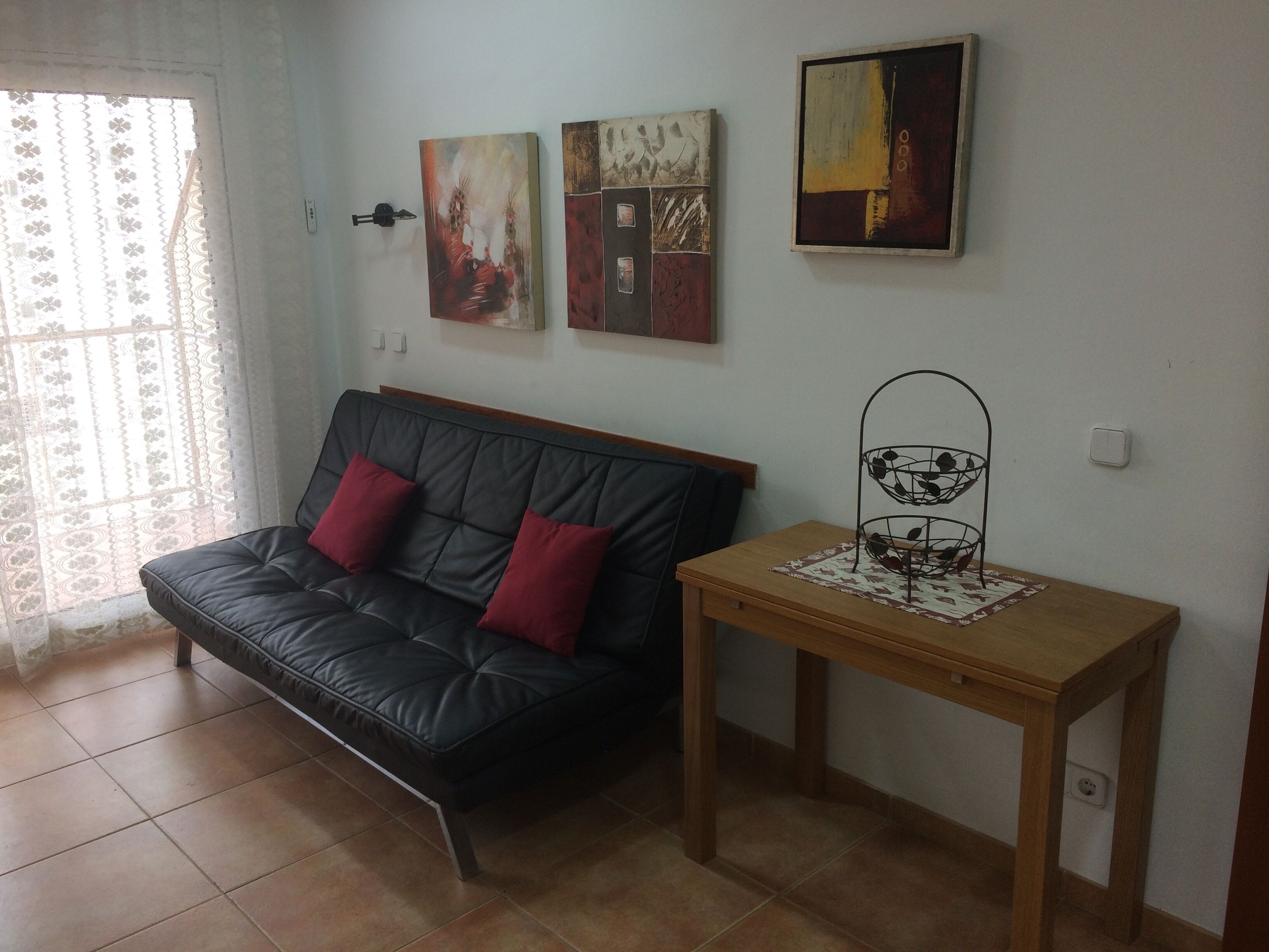 Un salón con un sofá de cuero negro, dos cojines rojos, una mesita de madera con una cesta de metal y tres obras de arte colgadas en la pared.
