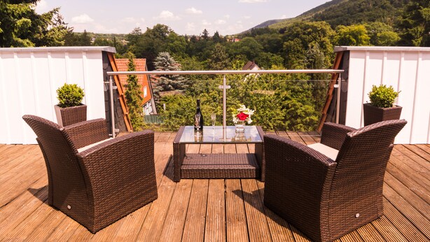 Eine Holzterrasse mit Korbmöbeln und einem Glastisch, mit Blick auf einen bewaldeten Hügel.