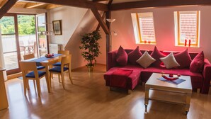Ein gemütliches Wohnzimmer mit einem roten Sofa, einem Holzesstisch und Stühlen. Eine Topfpflanze und eine Lampe sind ebenfalls vorhanden.
