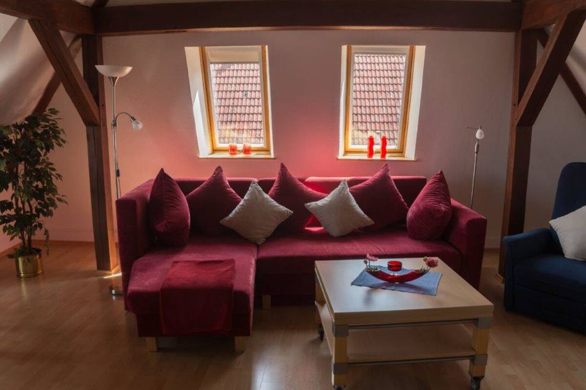 Une pièce chaleureuse avec un canapé rouge, un fauteuil bleu et une table basse en bois. Il y a deux fenêtres avec des cadres en bois et une plante dans le coin.