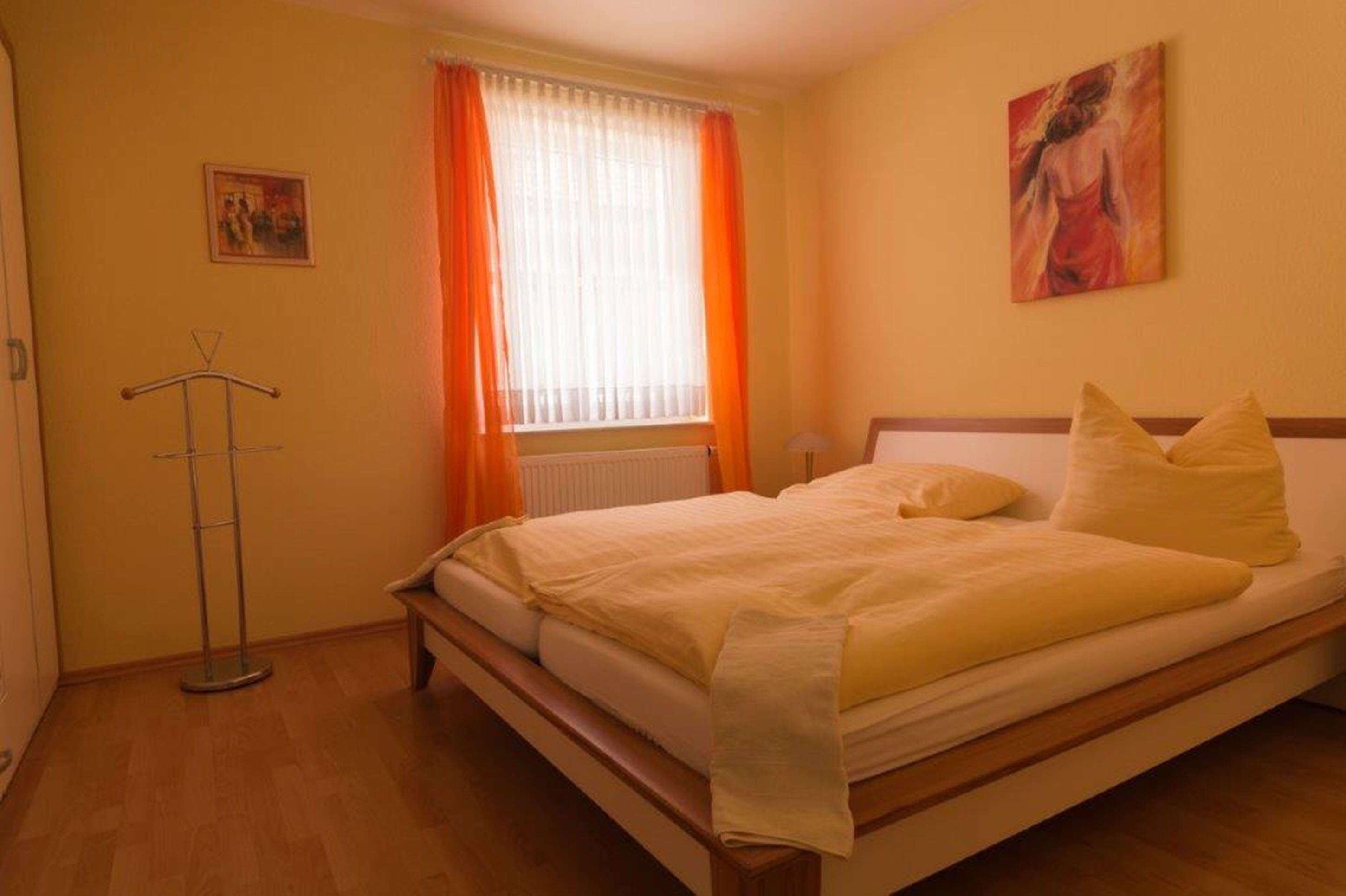 A bedroom with a bed, a window with orange curtains, a radiator, and a painting on the wall.