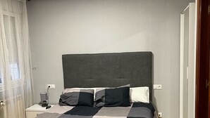 Cama bem arrumada com cabeceira cinza, mesa de cabeceira com abajur e tomada na parede.