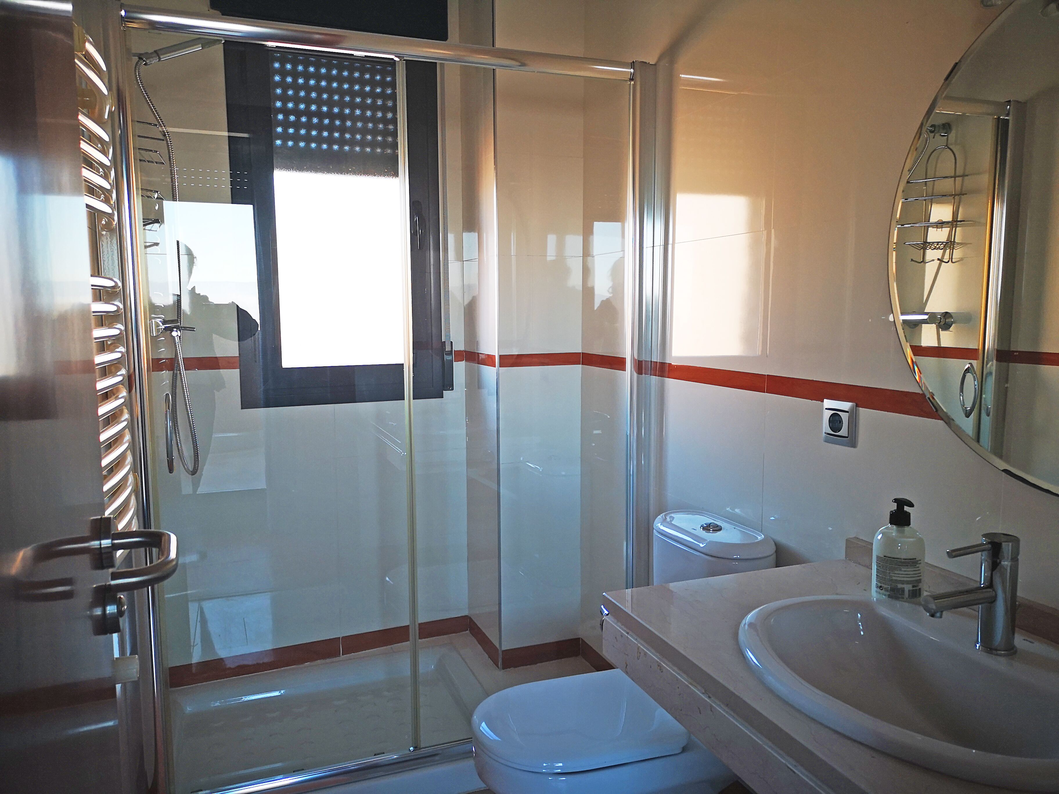 A bathroom with a glass shower enclosure, a toilet, a sink with a mirror, and a red stripe on the wall.