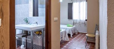 Una habitación con suelo de baldosas, una cama con cubrecama blanco, una mesita con un objeto verde y una silla.