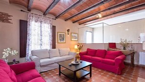 Una sala de estar con un sofá blanco, un sofá rojo y una mesita de centro. Hay dos cuadros enmarcados en la pared, una ventana con cortinas y un radiador.
