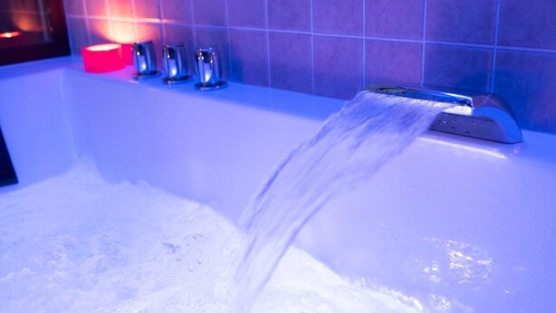 Una bañera con agua corriente, una vela roja y tres tazas metálicas sobre una pared de azulejos.