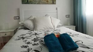 Habitación de hotel con cama, almohadas blancas, colcha con motivos florales, un cuadro de una playa y una cortina azul.
